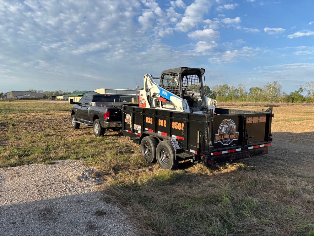 Black trailer with skid steer loader parked next to a pickup truck on an open field.