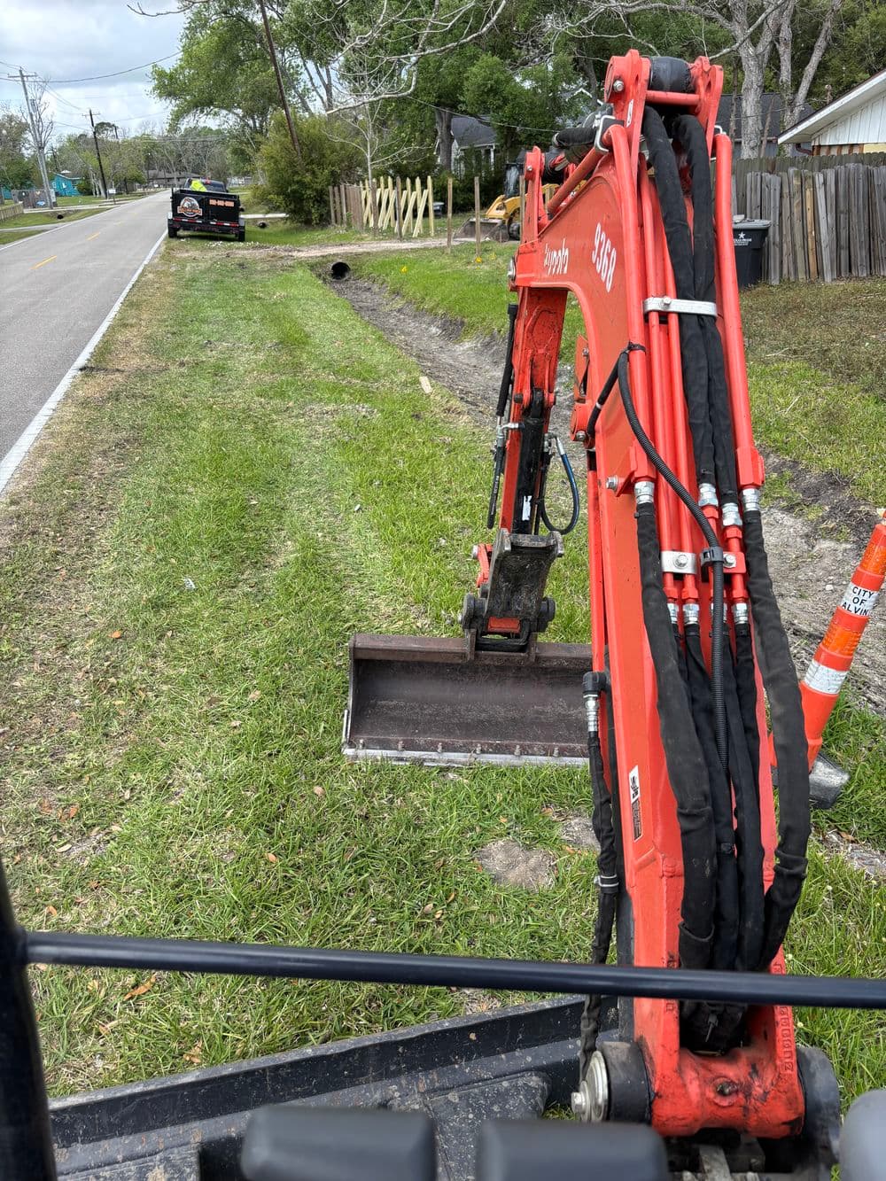 Excavator on a roadside digging near a drainage pipe with construction vehicle in background.
