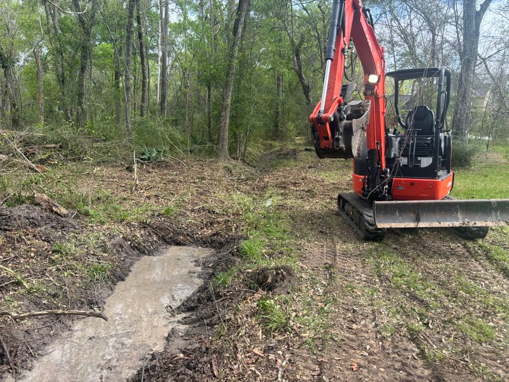 Mini excavator clearing a forested area, with water runoff and disturbed soil visible.