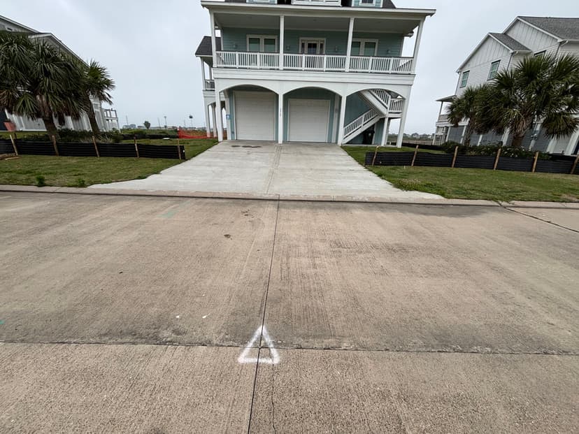 Driveway leading to a two-story beach house with palm trees and gray sky background.