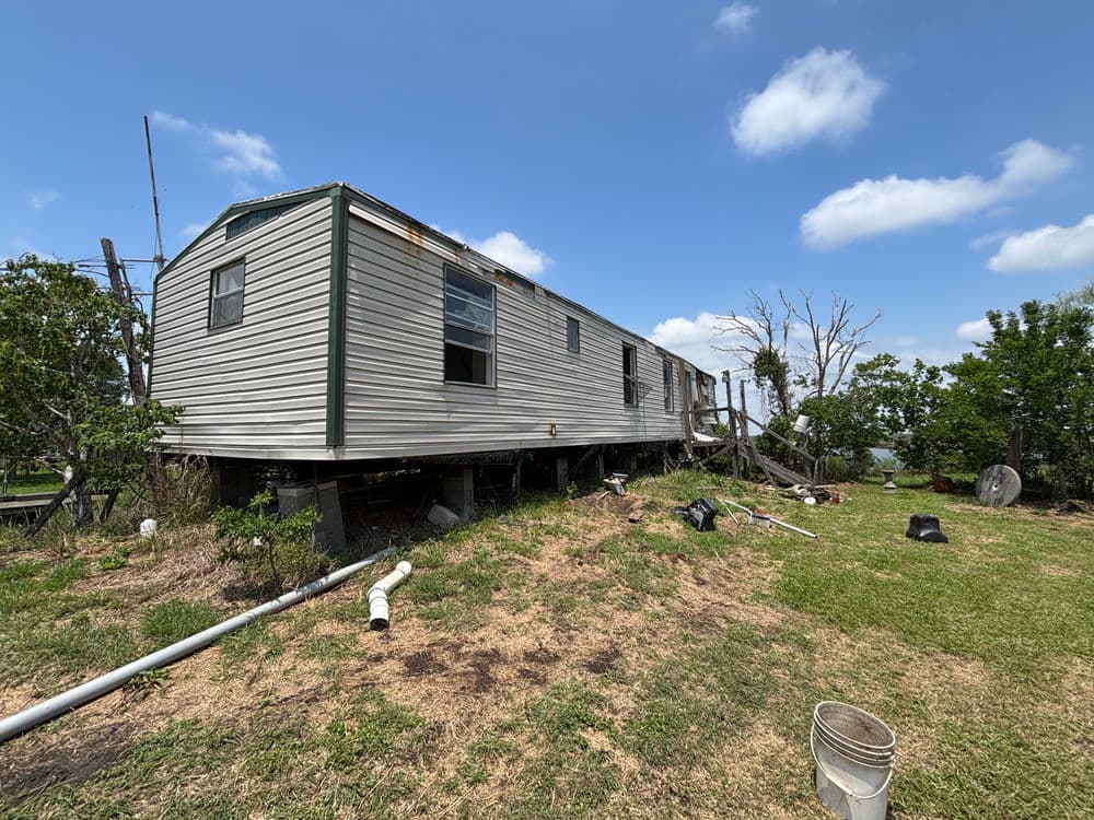 Abandoned mobile home on stilts surrounded by overgrown grass and scattered debris.