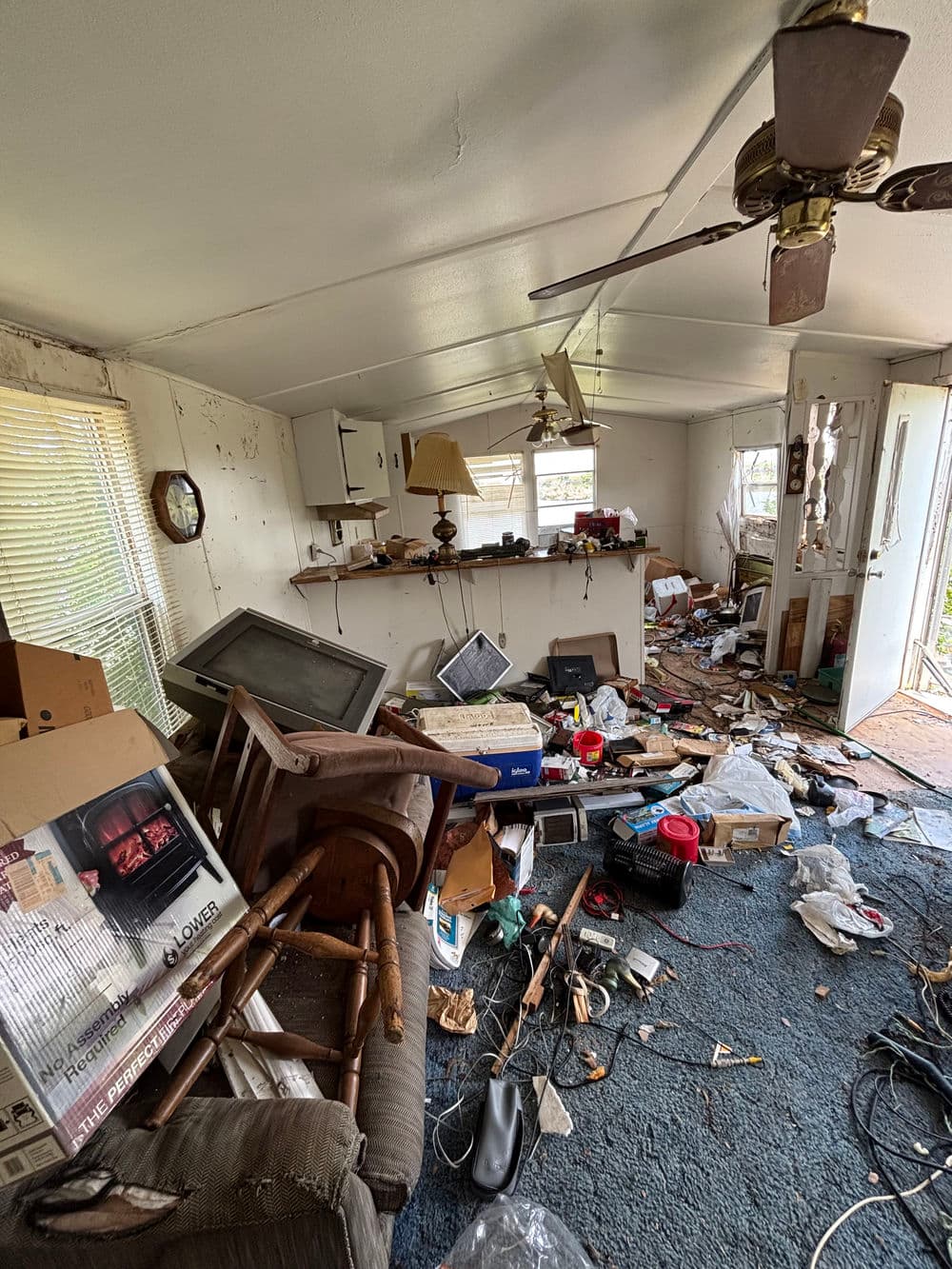 Cluttered living room with disorganized furniture, boxes, and debris on the floor.