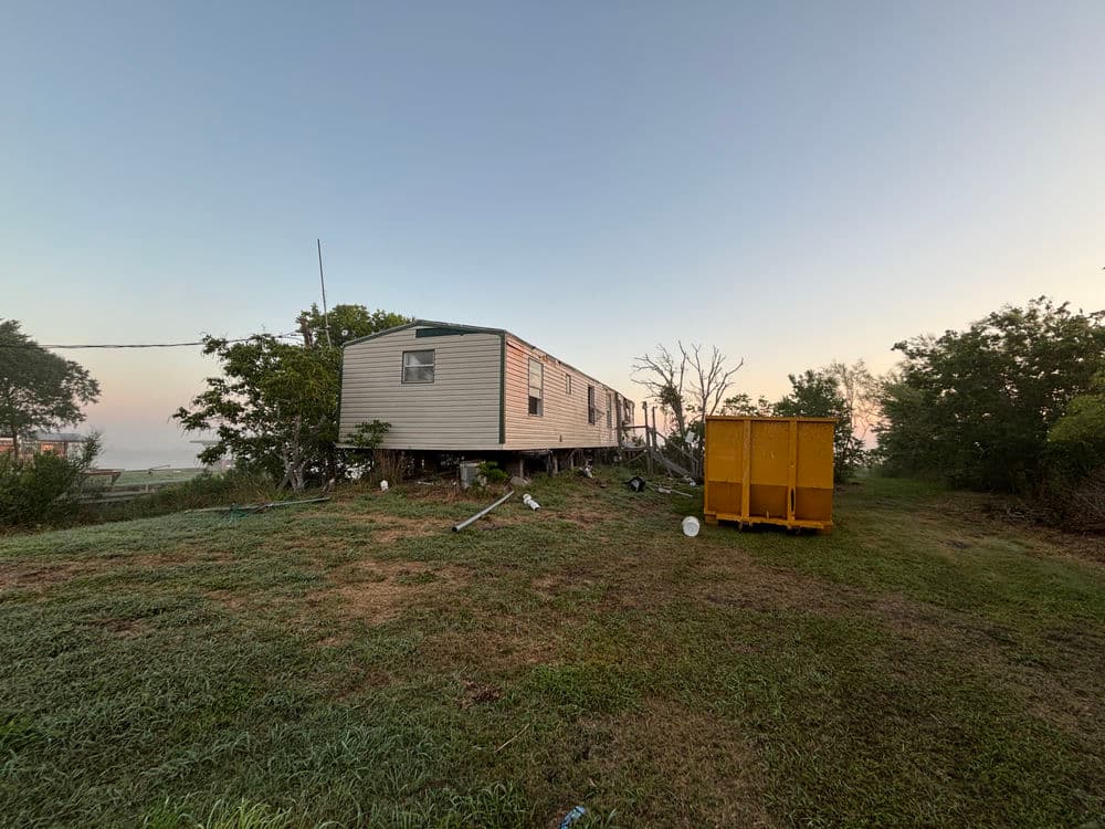 Single wide mobile home on stilts beside a yellow dumpster in a grassy area at sunrise.