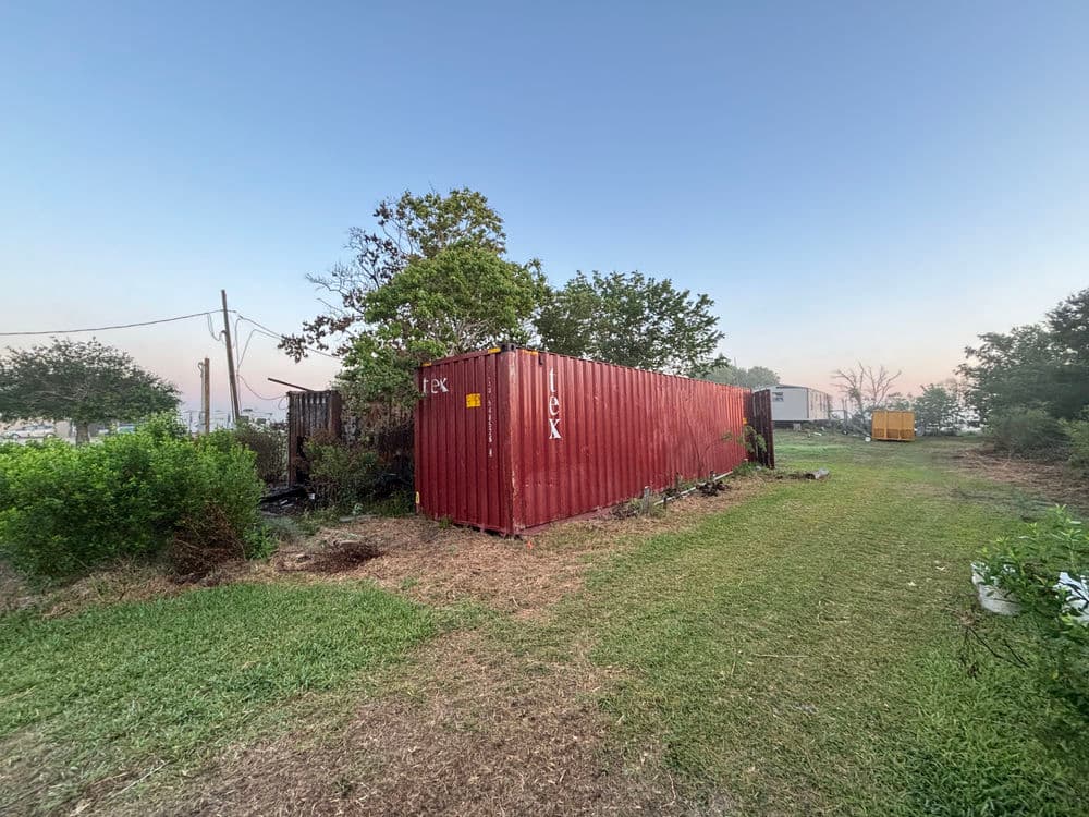 Red shipping container surrounded by greenery and trees at a rural location.