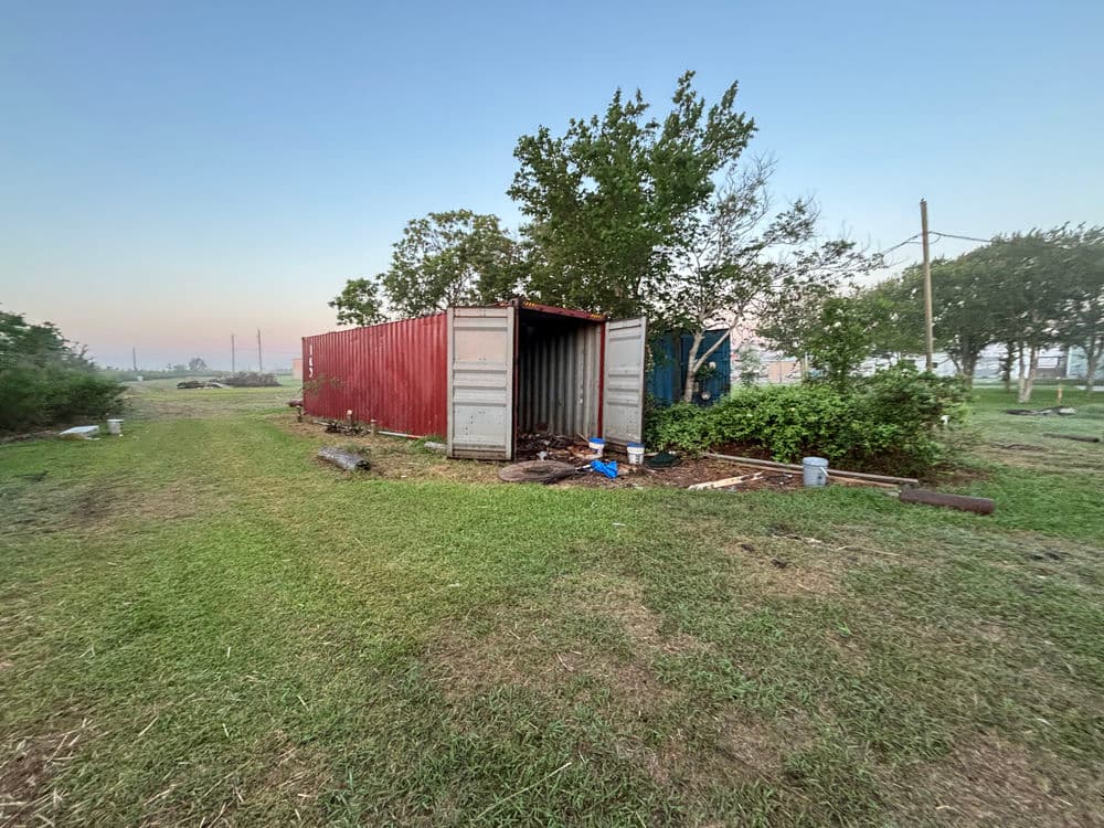 Red shipping container on a grassy plot surrounded by trees and utility poles at dawn.