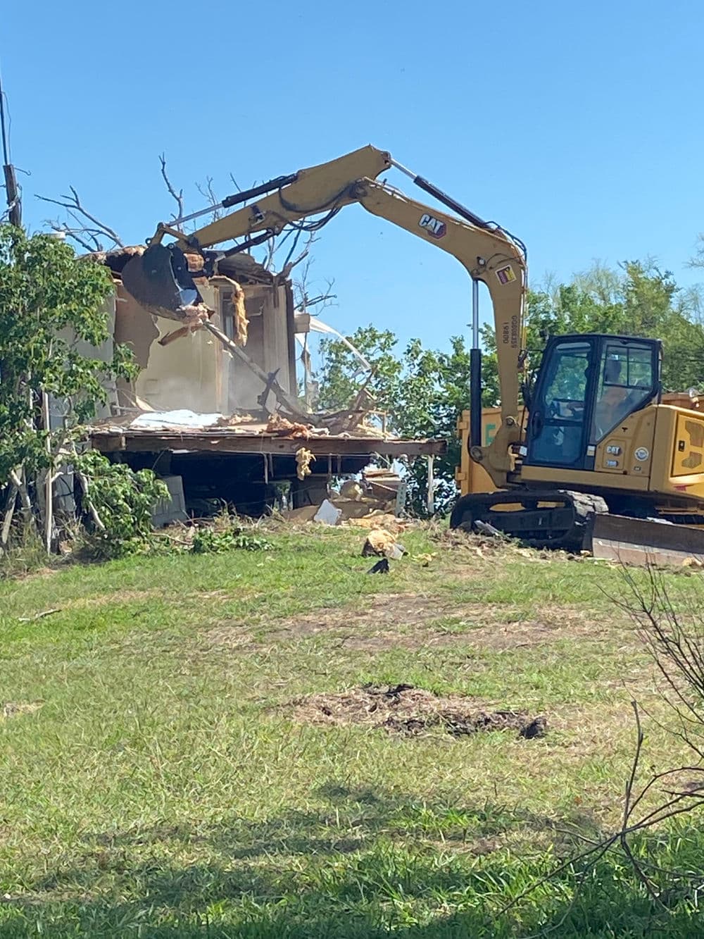 Excavator demolishing a building amidst clear blue skies and greenery.