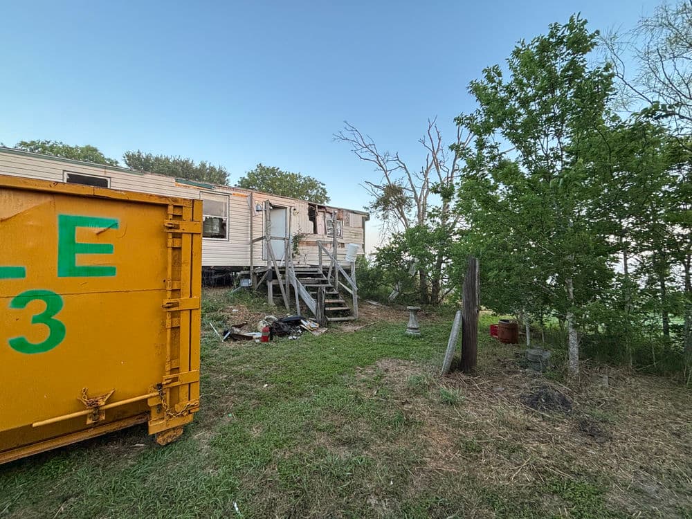 Old mobile home with steps, yellow dumpster, and overgrown greenery in rural setting.