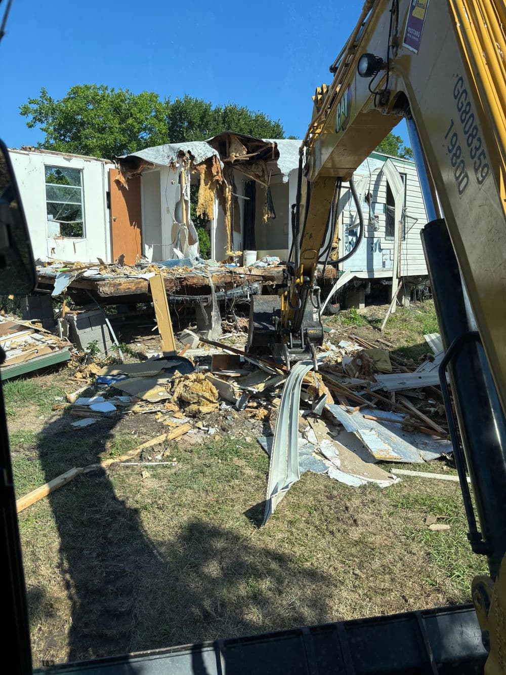 Excavator demolishing a house, debris scattered across the yard in bright daylight.