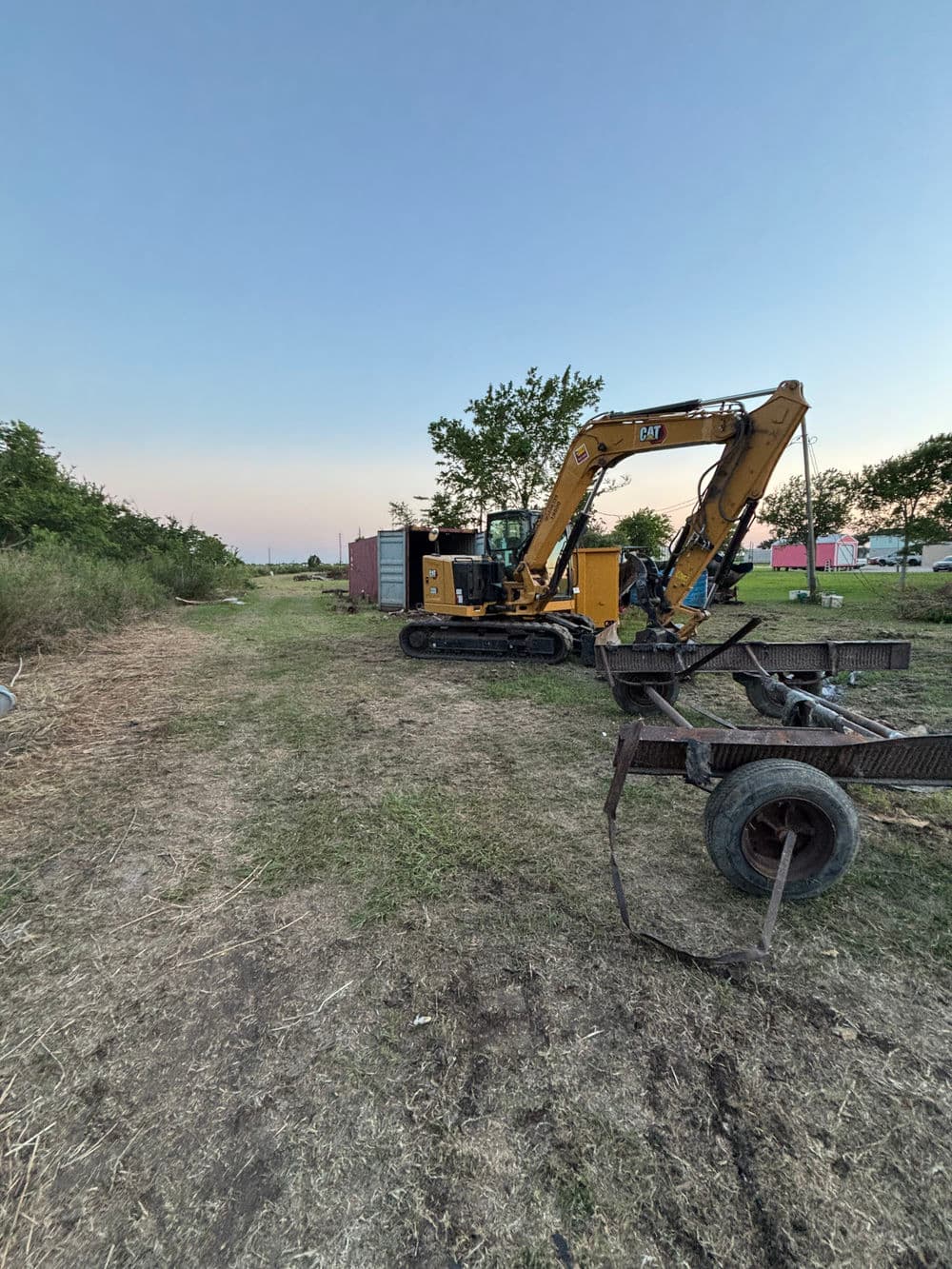 Excavator on a construction site with trees and container in the background at sunset.