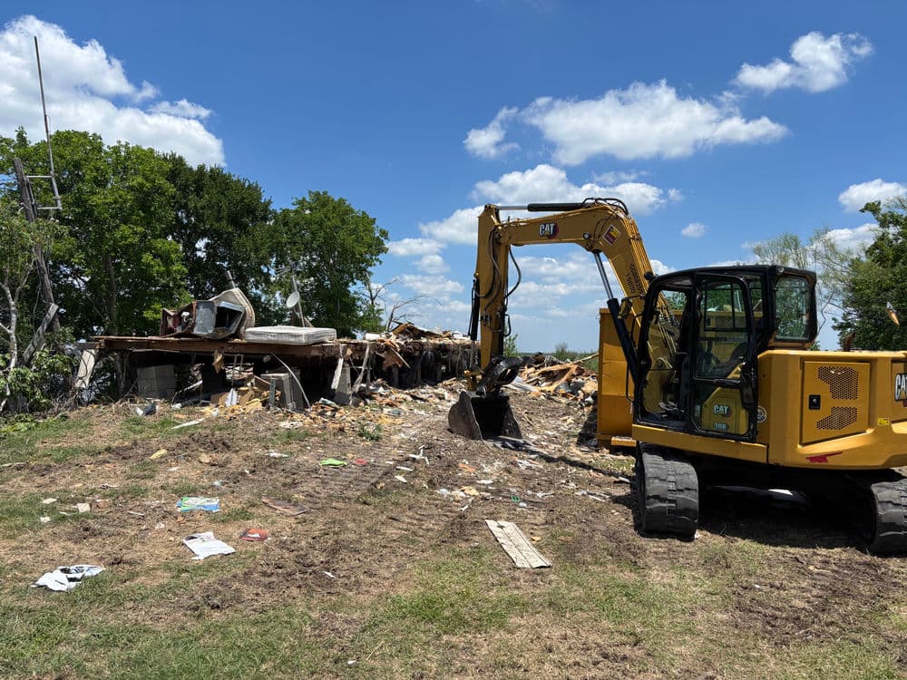 Excavator demolishing a destroyed building with debris under a blue sky.