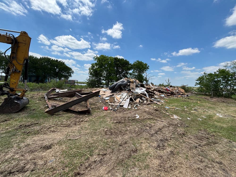 Construction debris pile with equipment under a blue sky and scattered trees in the background.