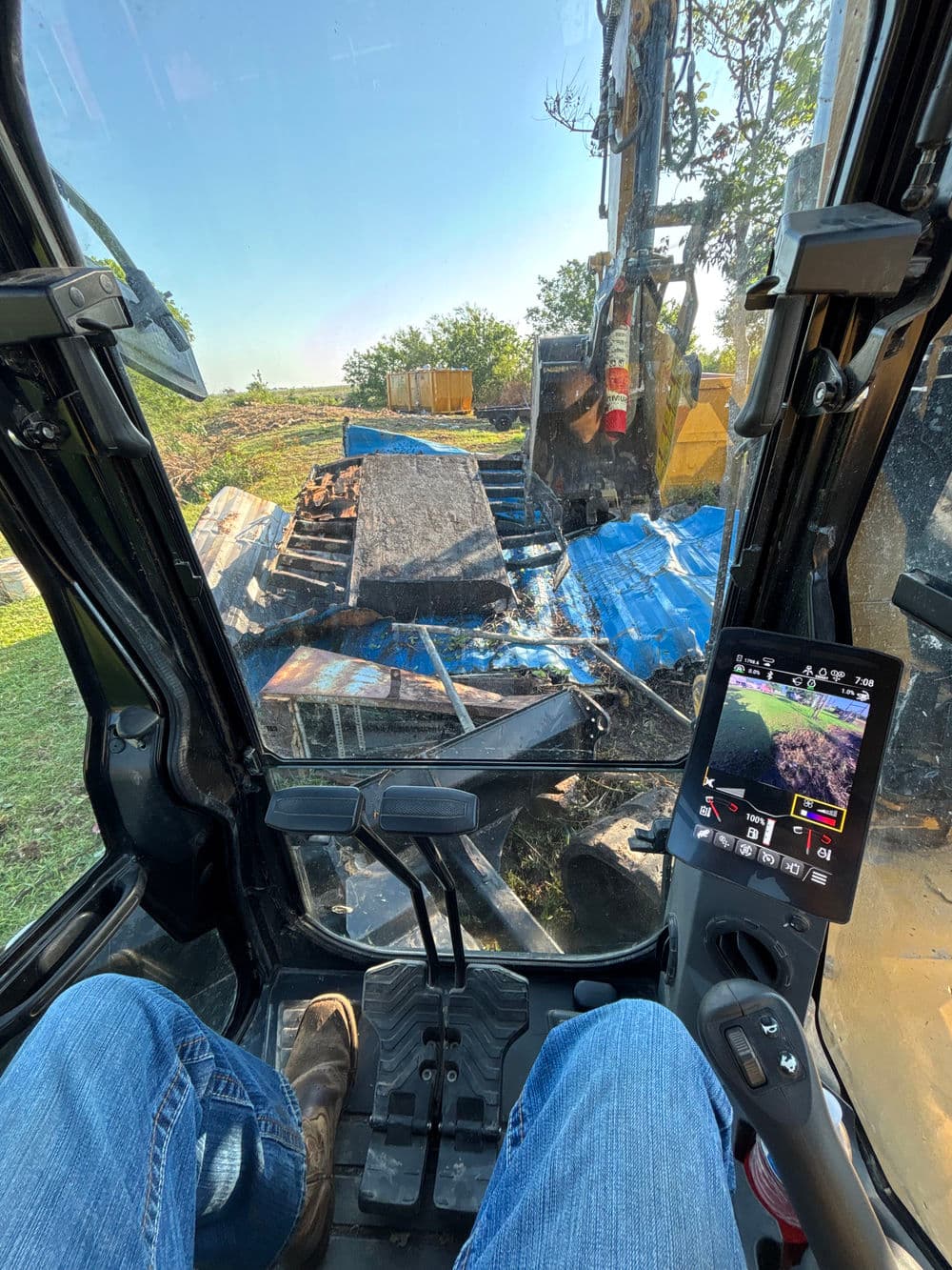 Operator's view inside a backhoe loader, with debris and blue tarps visible on site.