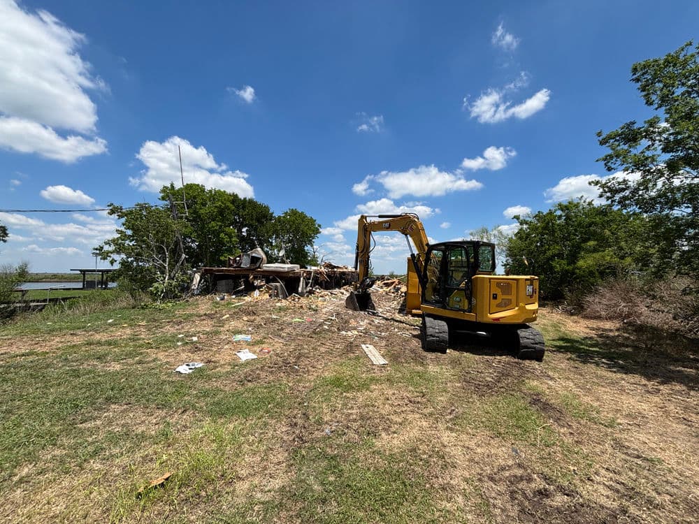 Excavator clearing debris from a demolished building under a blue sky with clouds.