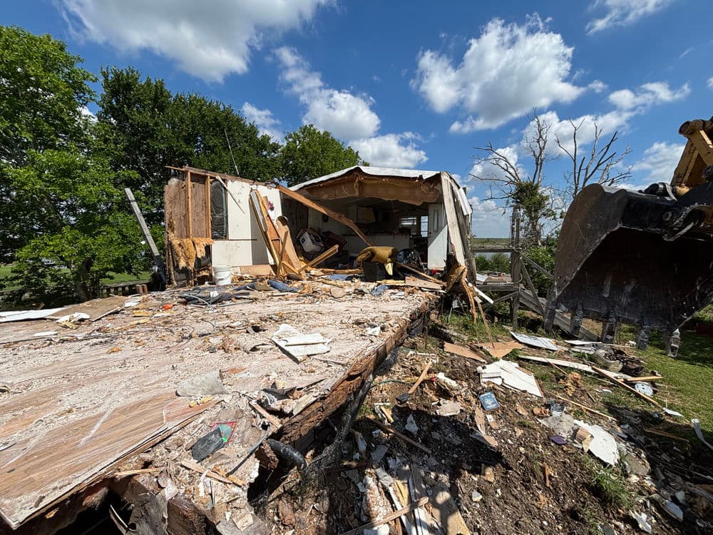 Damaged mobile home after storm, debris scattered, with construction equipment nearby.