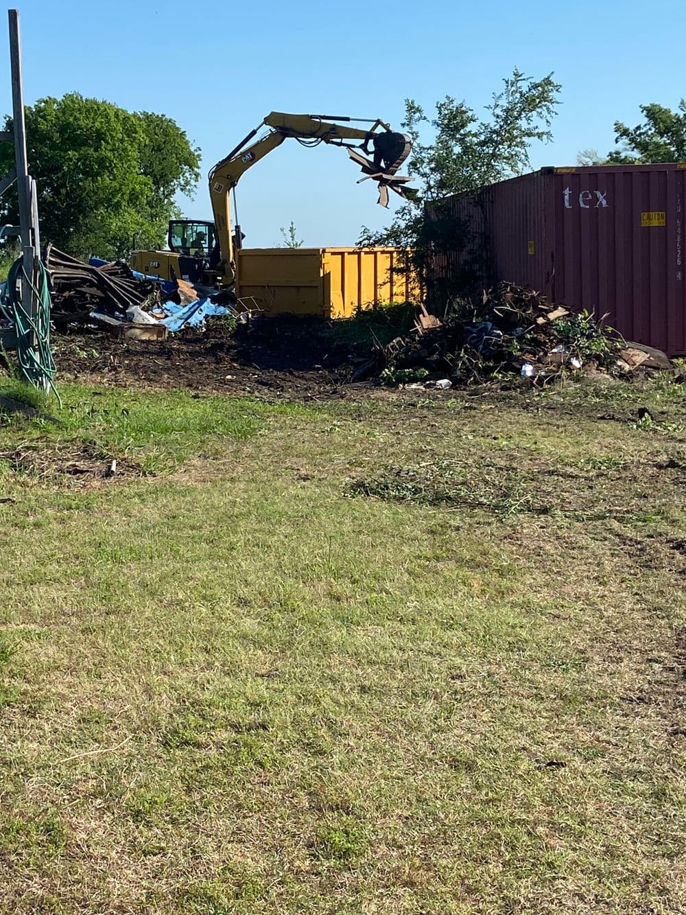 Excavator loading debris into a yellow dumpster at a clean-up site with shipping container.