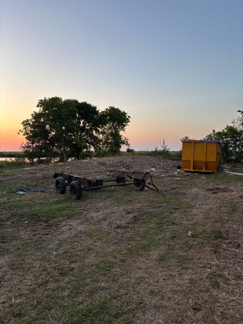Abandoned trailer chassis near a yellow dumpster at sunset in an open field.
