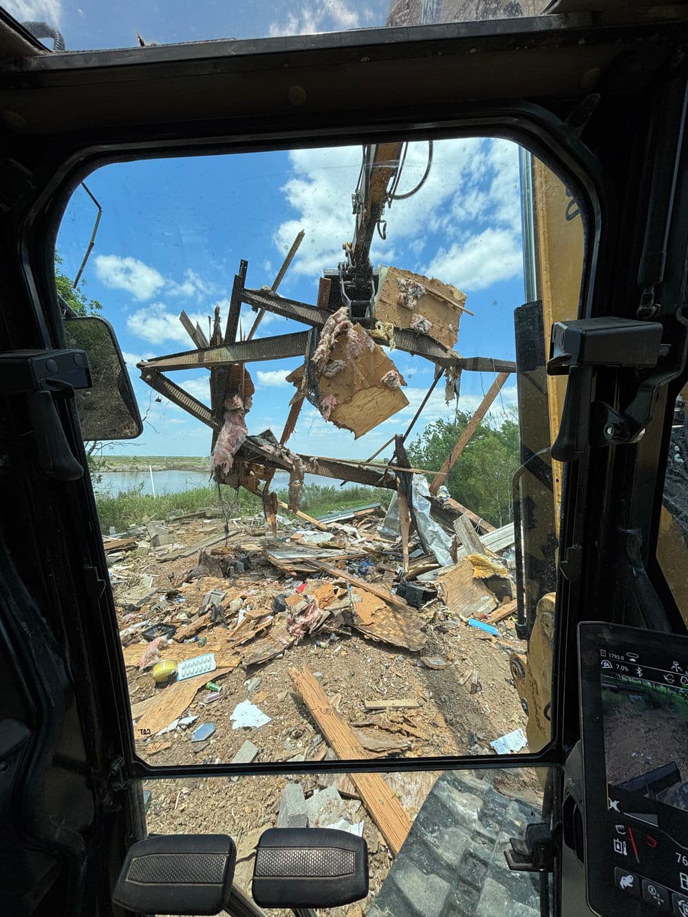 Excavator view demolishing a wooden structure amidst debris and blue sky.