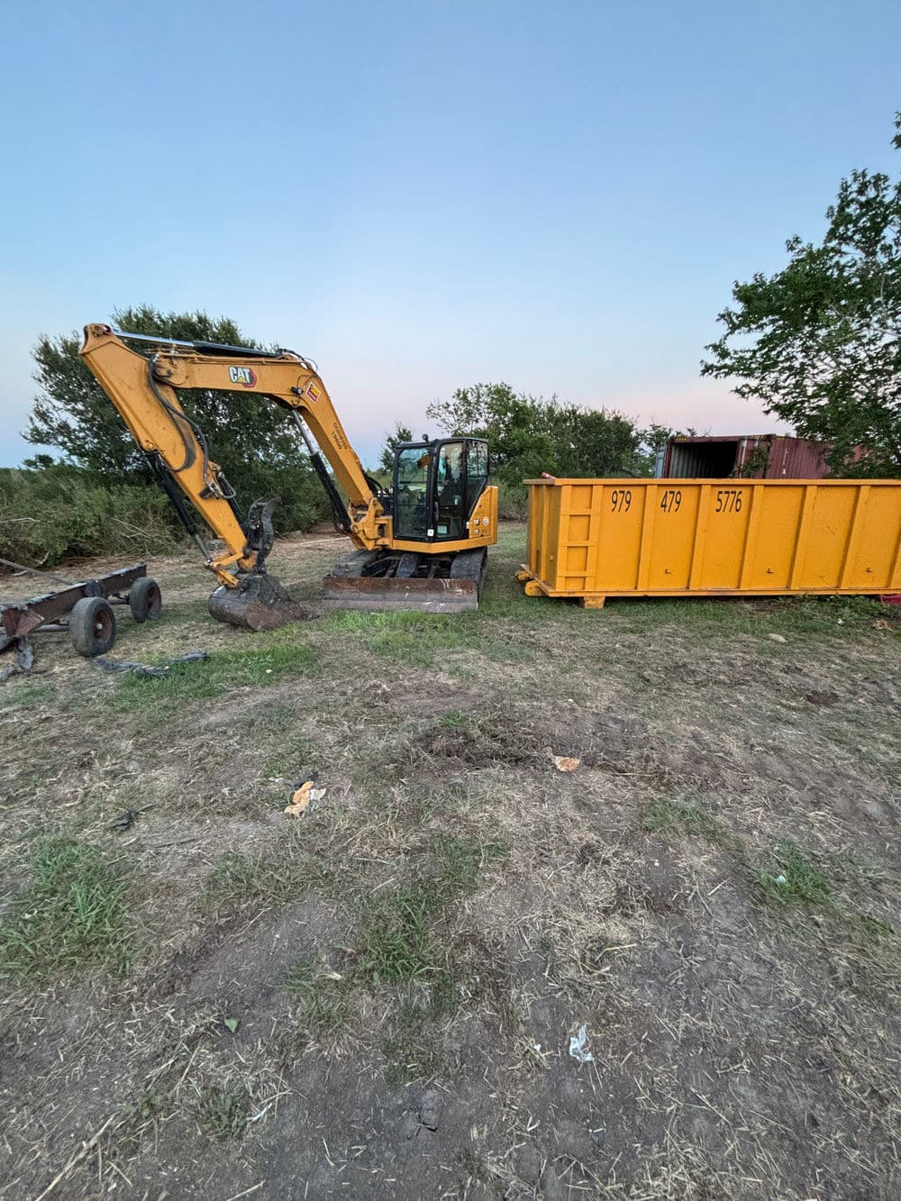 Caterpillar excavator working near a yellow dumpster on a construction site at sunset.