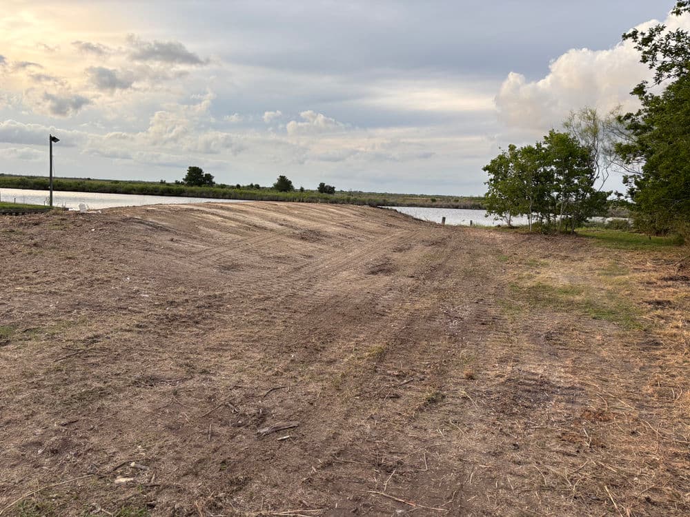 Dirt levee by a calm waterway under a cloudy sky, surrounded by sparse vegetation.