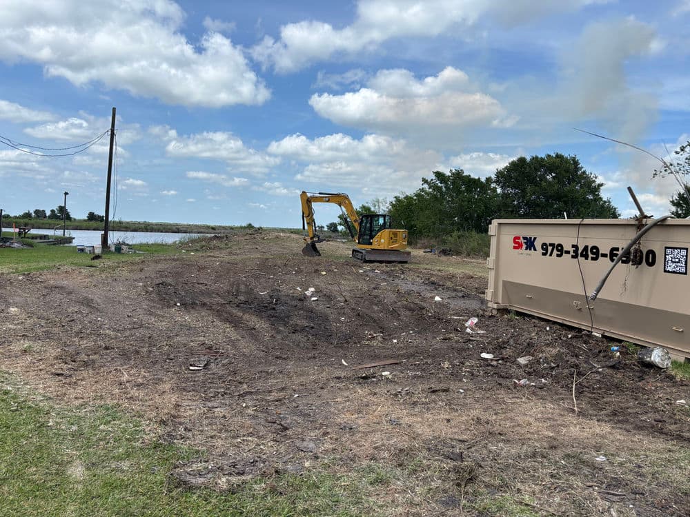 Excavator working on a cleared area near a body of water with debris and clouds in the sky.