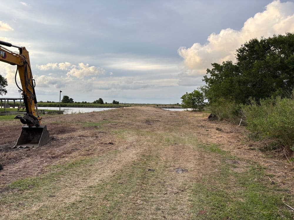 Excavator by a waterway with a cleared path and cloudy sky in the background.