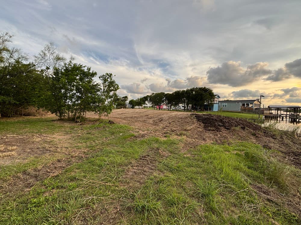 Cleared land by the water with trees and homes under a cloudy sky. Ideal for development.