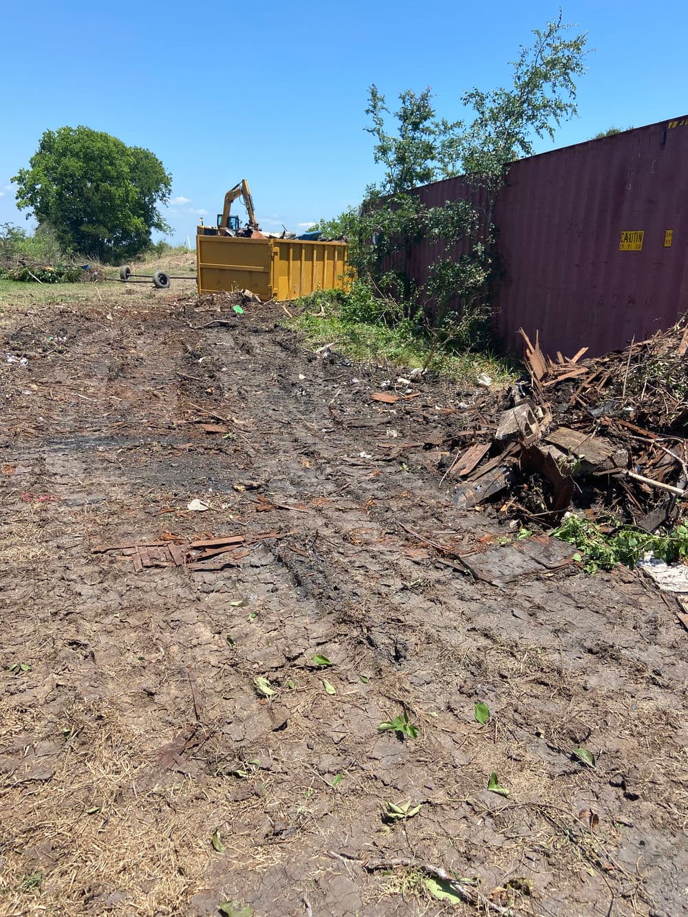 Construction site with a yellow dumpster, debris, and muddy ground under a clear blue sky.