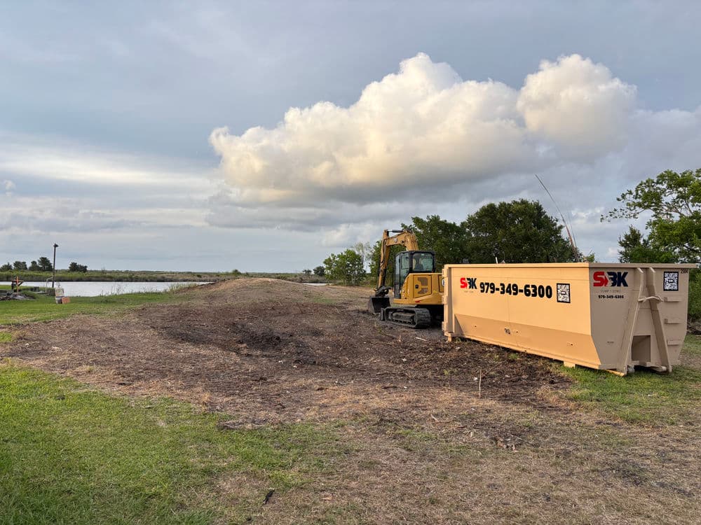 Excavator on a construction site near a water body with a large debris dumpster.
