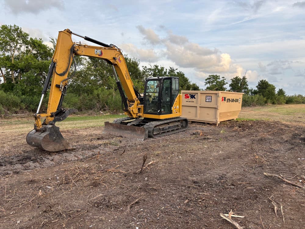 Excavator on a construction site with a dumpster in the background under a cloudy sky.