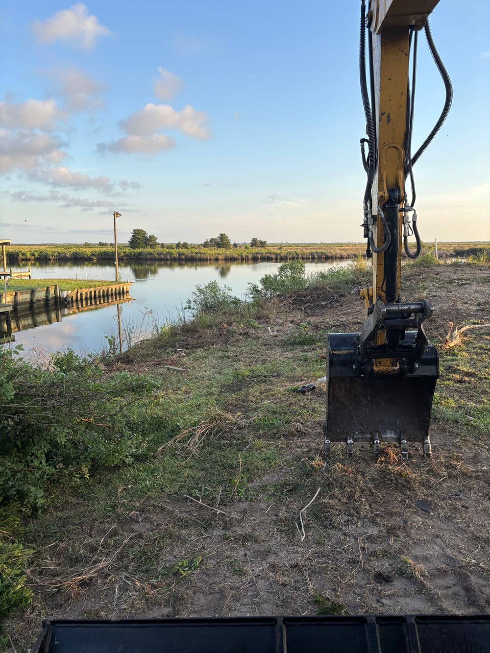 Excavator arm near a calm lake at sunset with lush greenery and a wooden pier.