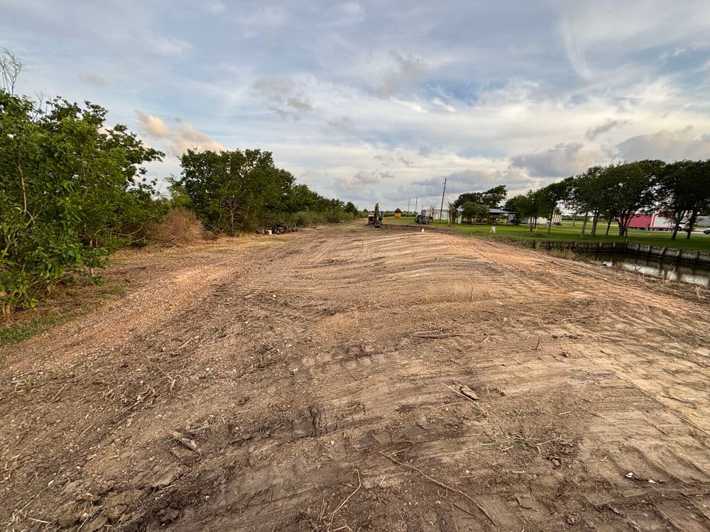 Cleared land along a canal with trees and equipment under a cloudy sky.