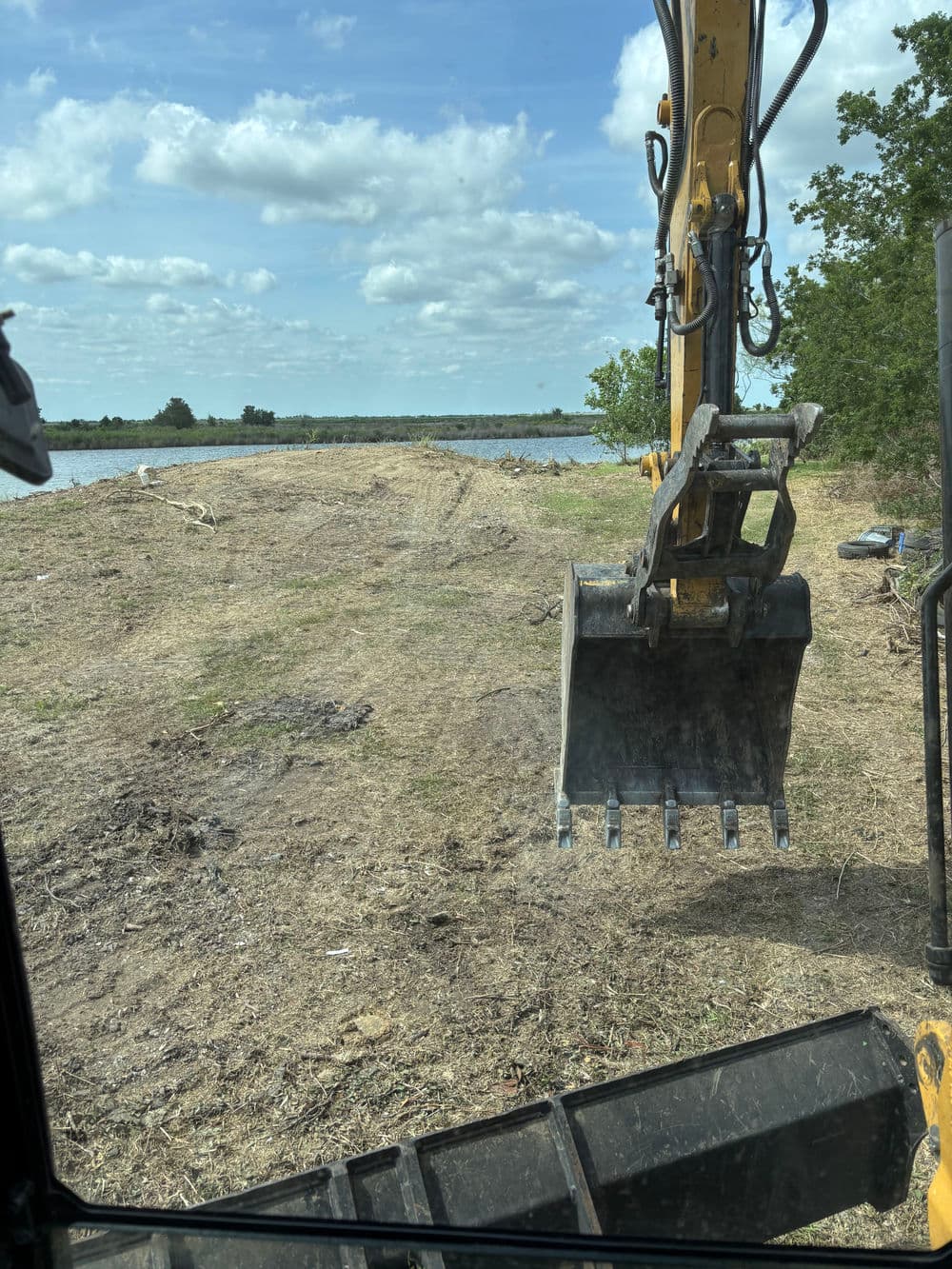 Excavator bucket over cleared land by a body of water under a partly cloudy sky.
