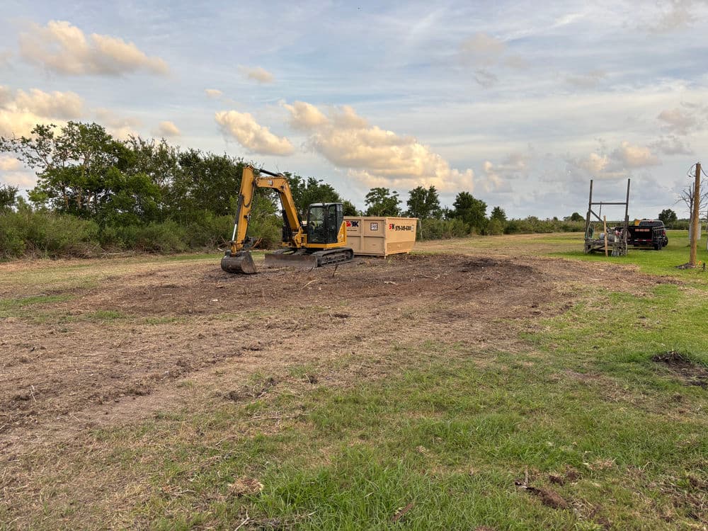 Excavator working on cleared land with trees and equipment in the background.