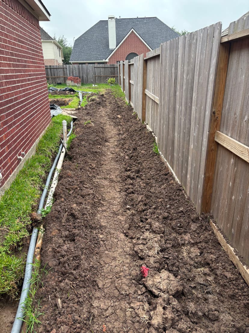 Dirt pathway beside a house, lined with wooden fence and utility pipes.